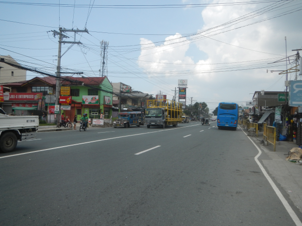 Eine belebte Stadtstraße mit Fahrzeugen, Radfahrern, Gebäuden, Strommasten, Schildern, Bäumen und einem bewölkten Himmel.