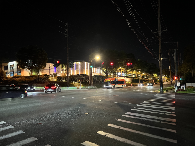 Eine nächtliche Stadtstraße mit fahrenden Autos und einem Bus, beleuchtet von Straßenlaternen und Ampeln, gesäumt von Bäumen, Gebäuden und Strommasten mit Drähten, unter einem dunklen Himmel.