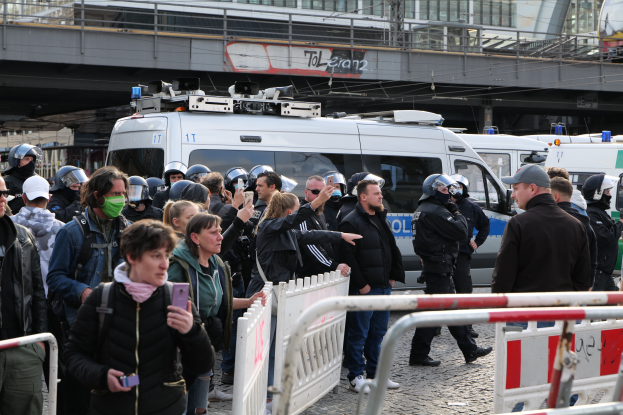Eine Gruppe von Menschen steht vor einer Reihe von Polizeiwagen, einige tragen Helme und halten Mobiltelefone, mit Absperrungen im Vordergrund und einer Brücke mit Geländern und Gebäuden im Hintergrund in Berlin, Deutschland.