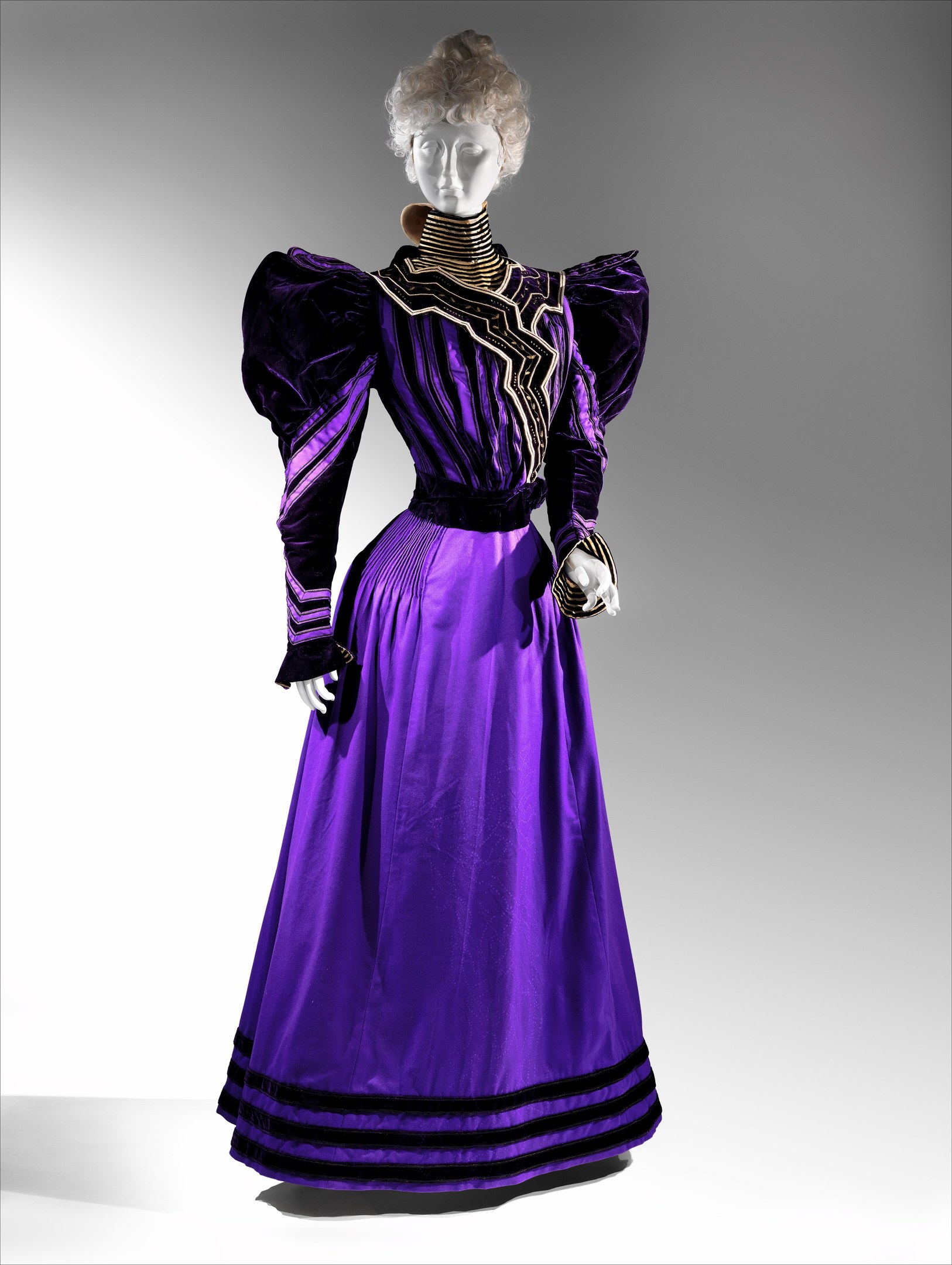 Eine Schaufensterpuppe in einem lila Abendkleid mit schwarzem und goldenem Muster, das eine eng anliegende Taille, einen langen Rock bis zum Boden mit goldener Stickerei und Perlen, eine Spitzenborte an der Taille, eine elegante Hochsteckfrisur und zarte Ohrringe aufweist.