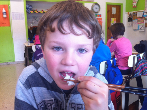 Ein Junge in einer grauen und schwarzen Jacke isst mit Stäbchen in einem Klassenzimmer, mit anderen Kindern hinter ihm, einer grünen und weißen Wand, Lehrplakaten an einer weißen Wand, einer braunen Tür und einer Wanduhr.