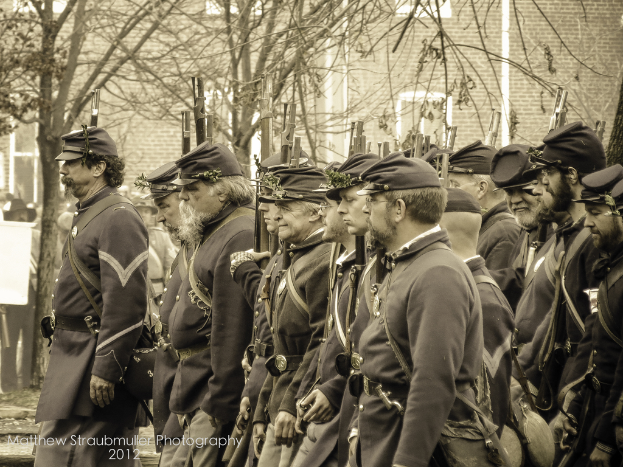 Mehrere Männer gehen auf der Straße mit Mützen und Waffen, mit Bäumen und einem Gebäude im Hintergrund; ein Wasserzeichen ist in der linken unteren Ecke sichtbar, schwarz-weißes Bild.