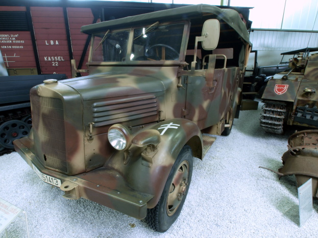 Alter Militärlastwagen in einem Museum ausgestellt, umgeben von anderen Fahrzeugen, mit Informationsschild links und Hintergrundobjekten an einer Wand.