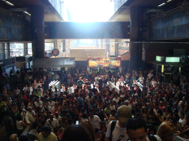 Eine Menschenmenge vor Ständen mit im Hintergrund fahrenden Fahrzeugen und Säulen an den Wänden.