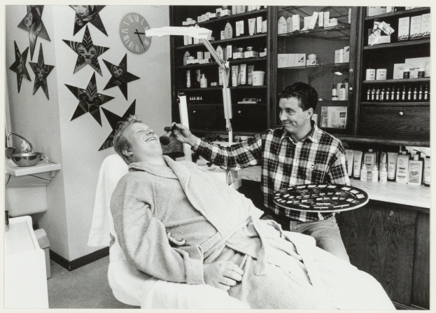 Ein Mann sitzt lachend in einem Zahnarztstuhl, während ein anderer Mann neben ihm steht und einen Teller hält, mit einem Waschbecken, Sternen, einer Uhr und Regalen voller Flaschen im Hintergrund.