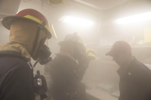 Feuerwehrleute in einem raucherfüllten Raum während einer Übung, einer hält eine Kamera, mit Ausrüstung und Beleuchtung im Hintergrund.
