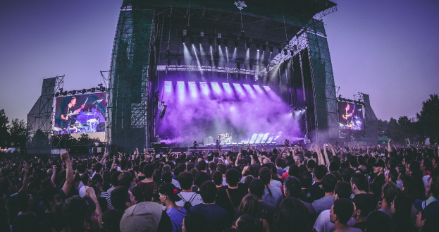 Eine große Menschenmenge bei einem Musikfestival schaut auf einer hell erleuchteten Bühne mit Instrumenten, Bildschirmen und einem Hintergrund aus Bäumen und einem klaren Himmel.