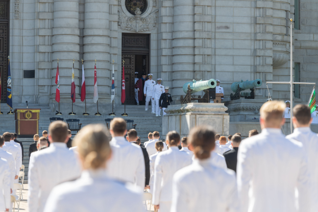 Gruppe von Menschen in weißen Marineuniformen vor einem Gebäude mit Säulen und Treppe bei einer Abschlusszeremonie mit Fahnen, Podium und Kanonen im Hintergrund.