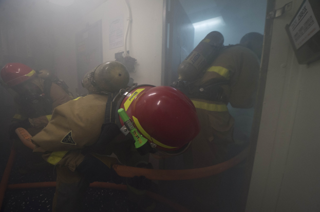 Feuerwehrleute in Schutzausrüstung betreten einen raucherfüllten Raum, mit Sauerstoffflaschen und Rohren sichtbar.