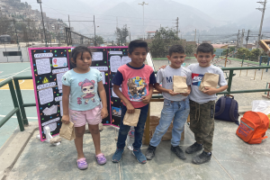 Gruppe von Kindern auf einem Basketballfeld, die Papierbeutel halten, mit Beuteln und Gegenständen rechts und einer Tafel mit Text links, vor einem Hintergrund aus Zaun, Pfosten, Straßenlaternen, Bäumen, Gebäuden, Hügeln und Himmel.