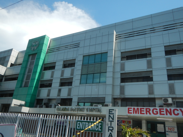 Ein großes Gebäude mit einer "Notfall"-Tafel, umgeben von einem Metallzaun, Pflanzen, Informationsschildern, Fahrzeugen und Drähten unter einem bewölkten Himmel.