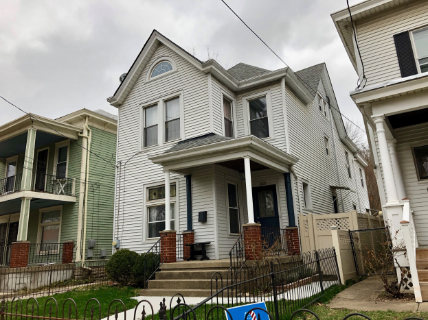 Reihe von Häusern mit Fenstern, Türen, Säulen, Geländern, Treppen und Dächern, umgeben von einem Metallzaun, Gras, Pflanzen, Bäumen und einem bewölkten Himmel, mit einem Mietschild am Boden, das die Adresse und die Details der Immobilie anzeigt.