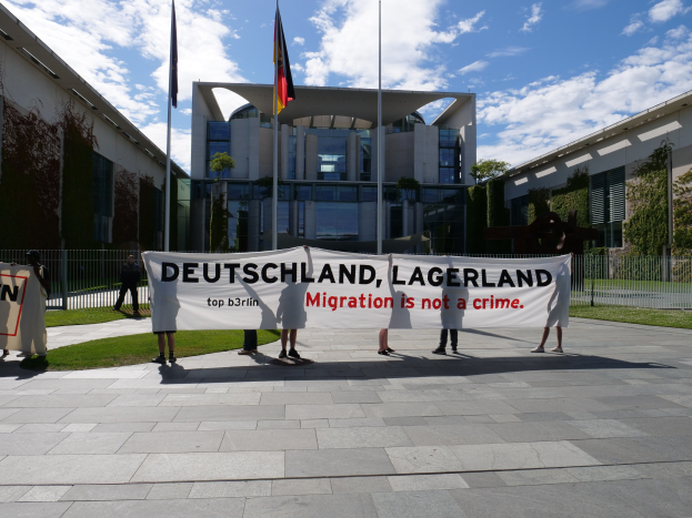 Eine Gruppe von Menschen hält ein Transparent mit der Aufschrift "Deutschland, Lagerland Migration ist kein Verbrechen" vor einem umzäunten Gebäude mit Bäumen und Flaggen.