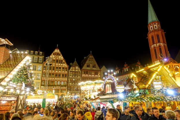 Ein geschäftiger Weihnachtsmarkt in Nürnberg, Deutschland mit Menschen um geschmückte Stände, Lichter, Gebäude mit Fenstern, ein Uhrenturm und einen dunklen Himmel.
