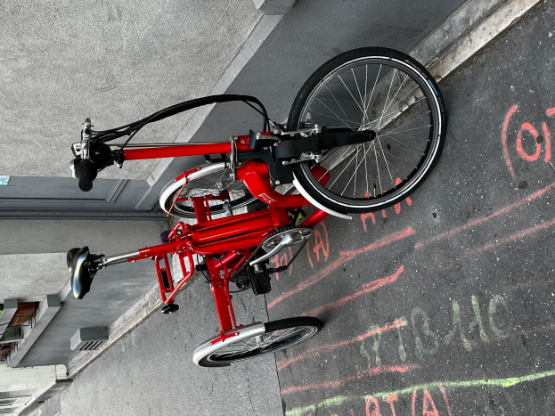 Ein rotes Klappfahrrad steht an der Seite einer Straße mit einem Gebäude im Hintergrund, und auf dem Fahrrad steht Text.