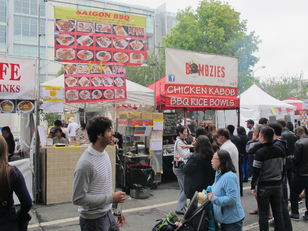 Menschen auf einer Straße vor der Stadt, mit einem Mann in einem grauen und weißen T-Shirt, der eine Wasserflasche hält, und einer Frau in einer blauen Jacke, die einen Kinderwagen schiebt, Stände mit weißen und roten Zelten, farbige Banner und Gebäude, Bäume und eine Straßenlaterne im Hintergrund.
