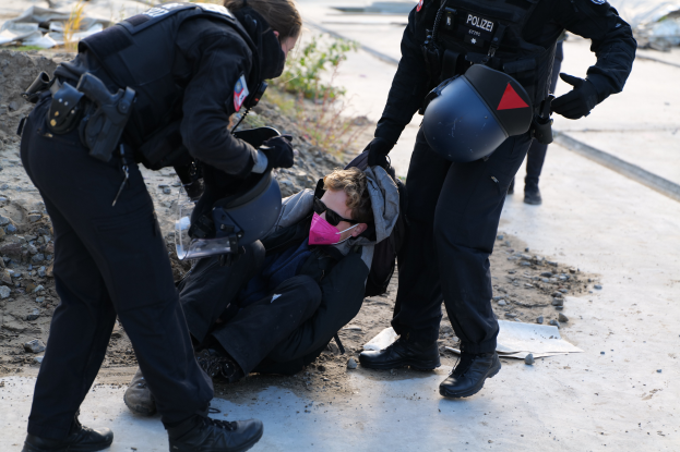 Zwei Polizisten in schwarzen Uniformen und Helmen tragen einen Mann von einer Szene weg, auf der Steine, Pflanzen und andere Gegenstände verstreut sind, mit einer Straße im Hintergrund.