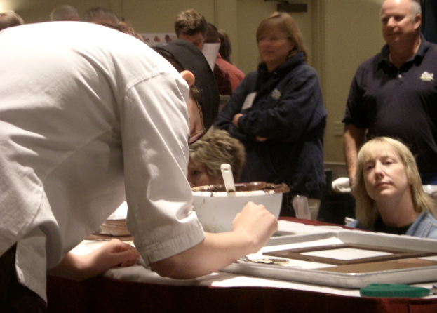 Eine Gruppe von Menschen steht vor einem Tisch mit einem Löffel, einer Schüssel und einem Tablett, mit einer Bibel und einer Wand im Hintergrund.