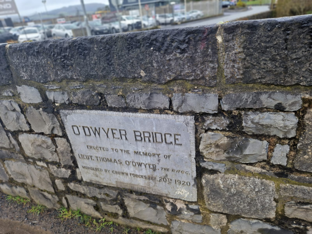Ein steinerner Wall mit einer Tafel, die "O'dwyer Bridge" trägt, vor einer Straße mit Fahrzeugen, Pfählen, Bäumen, Gebäuden und einem klaren blauen Himmel.