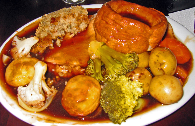 Weißer Teller mit Yorkshire-Pudding und Gemüse wie Karotten, Kartoffeln und Zwiebeln, umgeben von Gläsern, Papiertüchern und anderen Gegenständen auf einem Tisch.