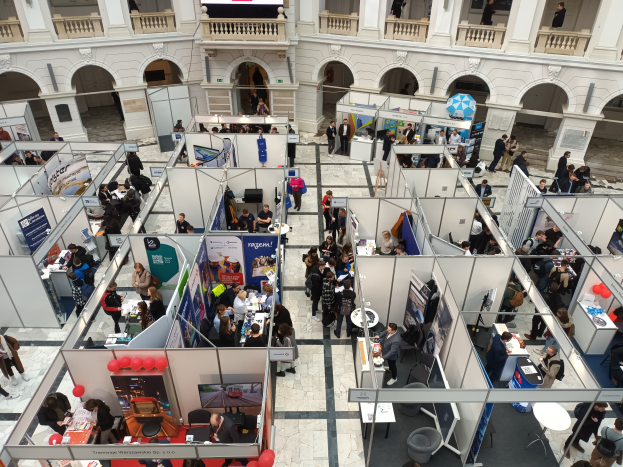 Eine große Gruppe von Menschen versammelte sich in einem Raum voller Stände, Banner, Tische, Stühle und Ballons, mit einem Gebäude im Hintergrund, das Bögen, Säulen, Geländer und einen Bildschirm aufwies.