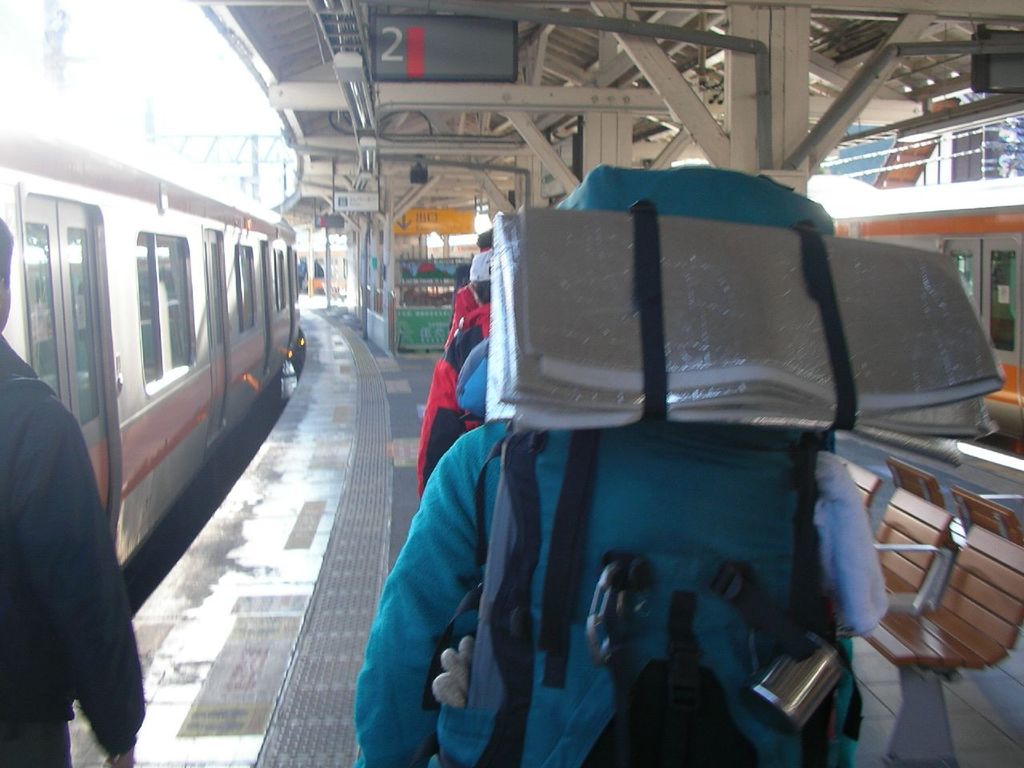 Ein Bahnhofsgebäude mit Menschen neben einem Zug auf der linken Seite, einem Stand in der Mitte, einem weiteren Zug auf der rechten Seite und einer Bank in der unteren rechten Ecke.