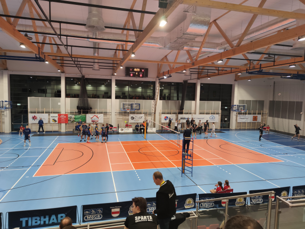 Eine Gruppe von Menschen, die in einer Turnhalle Handball spielen, umgeben von Zuschauern, die auf Stühlen und Stehplätzen sitzen, einer Leiter in der Mitte, Geländern und Texttafeln unten sowie Wänden, Säulen, einem Bildschirm und Deckenleuchten im Hintergrund.