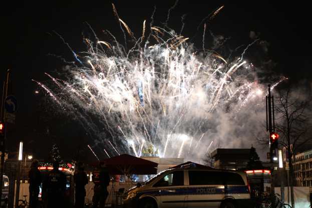Polizeiauto vor einer Menschenmenge während einer Silvesterfeier in Berlin geparkt, mit bunten Feuerwerken am Himmel.