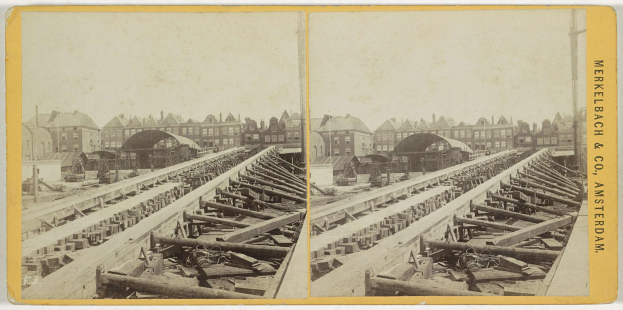 Schwarz-weiß-Foto einer Bahntrasse mit Gebäuden im Hintergrund, Text 'Merkelbach & Co, Co. Amsterdam' rechts und hölzernen Gegenständen im Vordergrund.