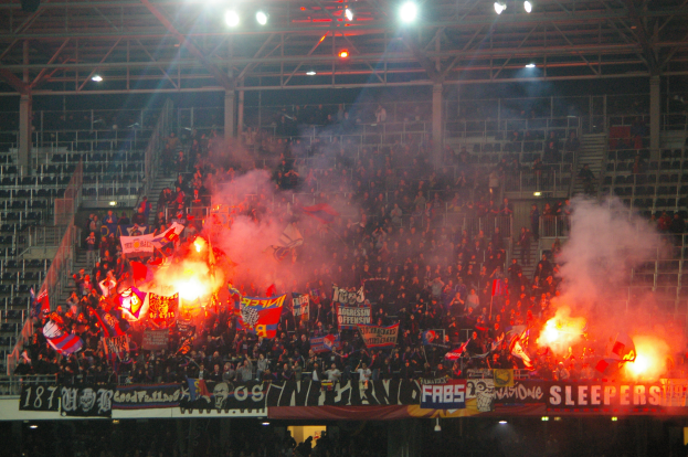 Eine große Menschenmenge in einem Stadion hält Fahnen und Banner, mit Rauchkerzen, unter einer Decke mit Deckenleuchten und Metallrahmen.
