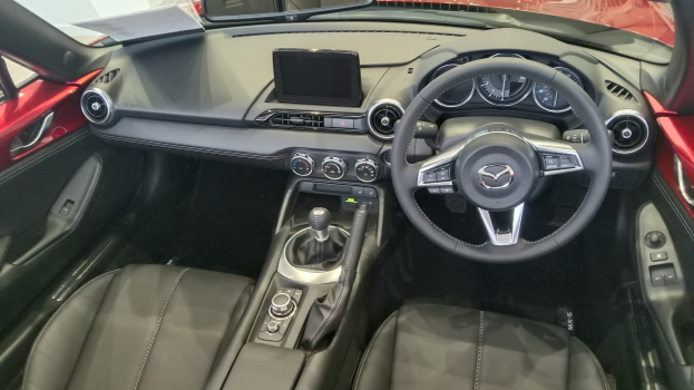 Innenansicht eines roten Mazda MX-5 Miata RF mit schwarzen Ledersitzen, Lenkrad, Schaltstange, Knöpfen, Bildschirm und Spiegel.