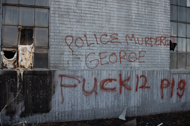 Ein Gebäude mit der Graffiti-Beschriftung "Polizei Morden George Fuck 12 P19" an der Seite, mit Fenstern und diversen Gegenständen vorne auf dem Boden.