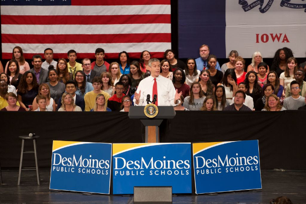 Ein Mann in einem weißen Hemd und roter Krawatte steht an einem Rednerpult mit drei Texttafeln, einem Hocker mit Mikrofon und einem Publikum im Hintergrund mit einer Fahne und Text an der Wand.