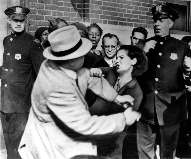 Zwei Personen kämpfen im Vordergrund, während eine Gruppe von Menschen, einige mit Mützen, im Hintergrund an einer Wand steht.