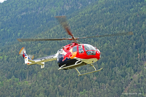 Ein roter Bullenhubschrauber fliegt über einen grünen Wald mit Bäumen auf den Hügeln im Hintergrund und einem klaren blauen Himmel darüber.