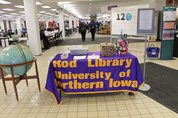 Eine Ausstellung mit einem Tisch, auf dem verschiedene Landesflaggen, eine Box, Papiere und ein Banner liegen, eine Kugel links daneben, ein Anschlagbrett rechts mit ein paar Leuten in der N√§he und mehreren Tischen und Stühlen im Hintergrund.