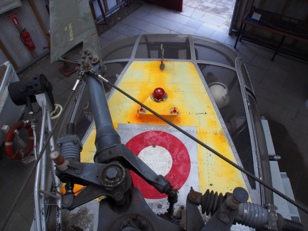 Nahaufnahme eines Hubschraubers auf einem Boden in einem Gebäude, mit einem Feuerlöscher links daneben und einer Bank rechts daneben, was Wartungsaktivitäten anzeigt.