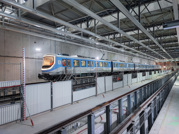 Ein blauer und weißer Zug fährt durch eine große Bahnhofshalle mit einer Plattform auf der rechten Seite und einer Wand im Hintergrund, unter einer Decke mit Stangen und Lampen, Teil eines neuen Schienensystems in Deutschland.