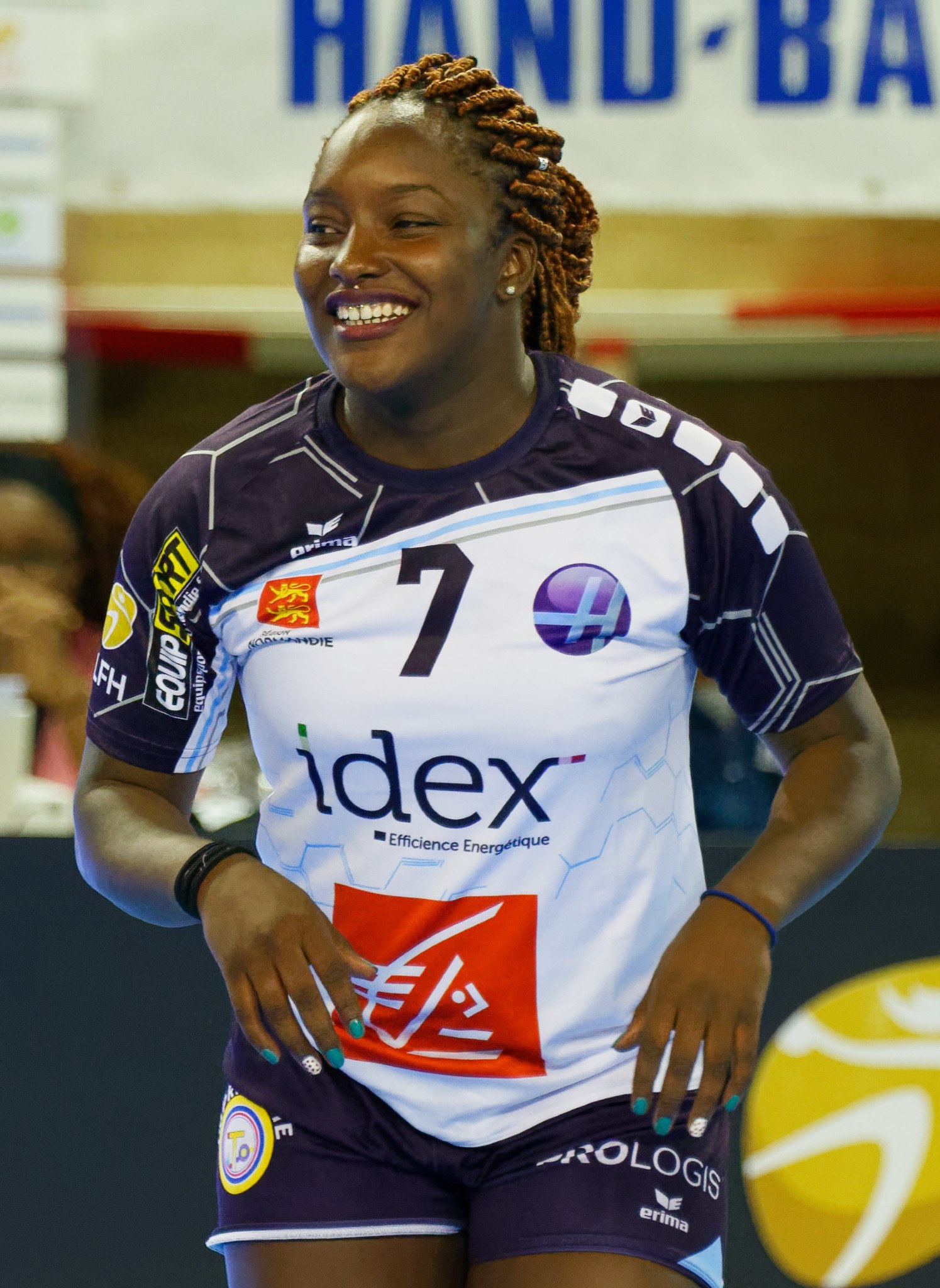 Professionelle Handballspielerin in lila-weißer Uniform, die auf einem Feld lächelt, mit einem FIVB-Handball-Weltmeisterschaft-2019-Schild im Hintergrund.