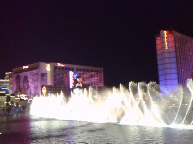 Ein Brunnen in der Mitte mit Wasser unten, umgeben von Gebäuden, Plakaten, Bildschirmen, Lichtern, Pflanzen und Himmel im Hintergrund.