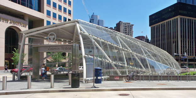Eine belebte Stadtstraße mit hohen Wolkenkratzern, eine Bushaltestelle mit Glasdach, Fußgänger, Fahrzeuge, ein Metallzaun, ein Müllcontainer, Strommasten, ein Schild, eine Ampel, Bäume, ein Baukran und ein bewölkter Himmel.