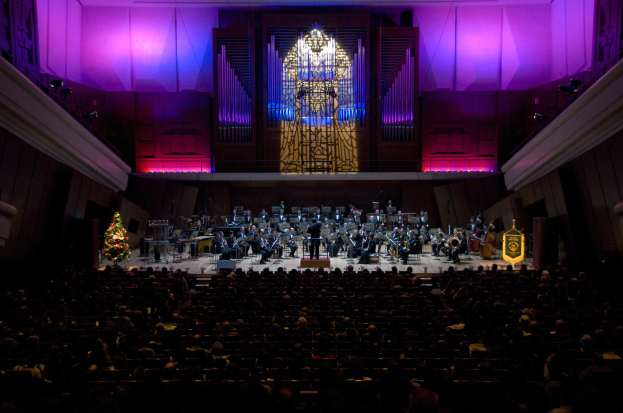 Großer Saal mit Menschen bei einem Konzert, Musiker auf der Bühne, ein Weihnachtsbaum im Hintergrund und geschmückte Wände mit Lichtern.