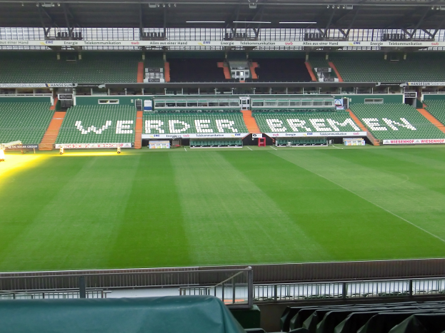 Ein großes Stadion mit einem grünen Feld, umgeben von Stühlen, Werbetafeln, Lampen und Stangen, mit Geländern und einem drapierten Stuhl unten.