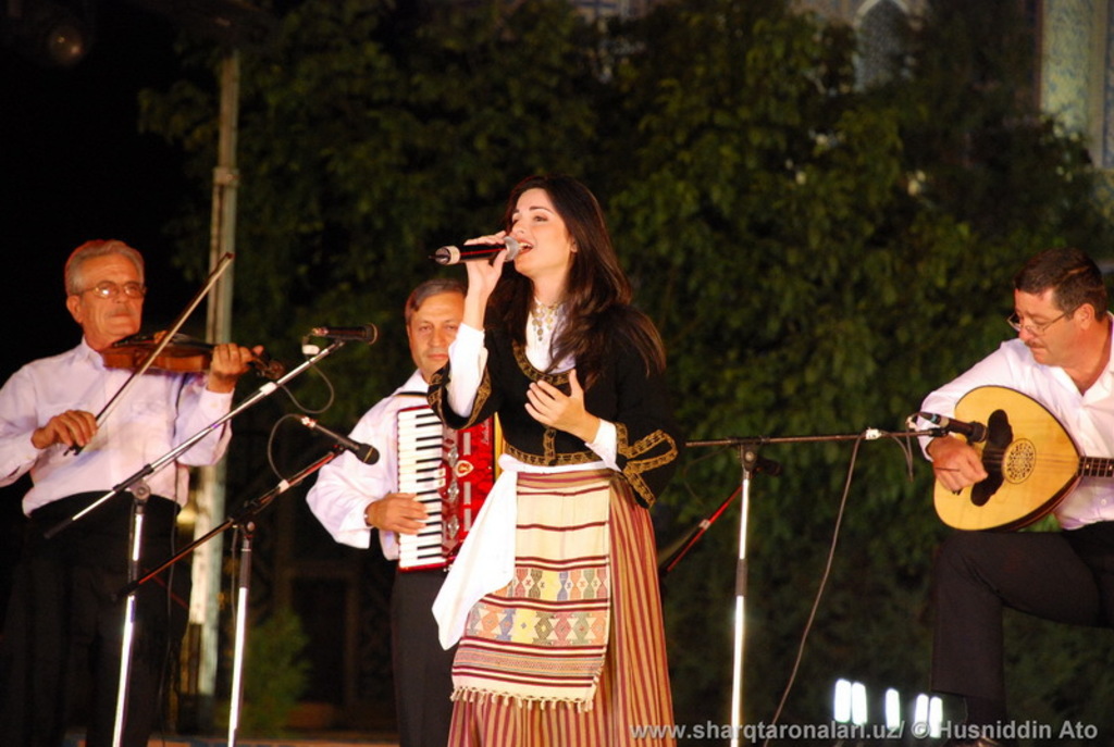 Eine Frau in einem Kleid singt in ein Mikrofon vorne, mit drei Männern, die Musikinstrumente vor Mikrofonen spielen, und Bäumen mit Lichtern im Hintergrund.