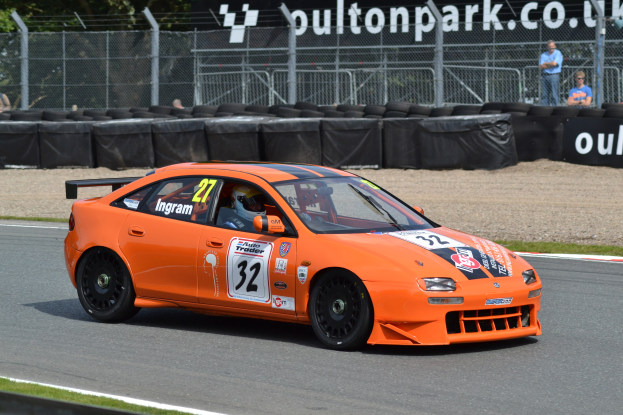 Ein oranges Rennauto mit einem Helmträger am Steuer rast eine Strecke entlang, mit Reifen, Absperrungen, Zuschauern, Bäumen und einem Banner im Hintergrund.