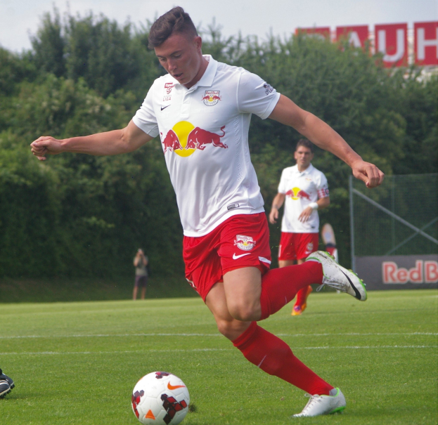 Ein professioneller Fußballspieler, der einen Fußball auf einem Rasenfeld mit Bäumen und einem klaren blauen Himmel im Hintergrund kickt, in der Nähe eines Schildes mit der Aufschrift "RB Leipzig."