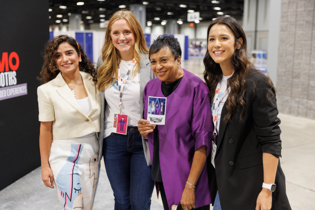Vier Frauen auf ID-Karten lächeln zusammen an einem Konferenzstand, mit einem Textschild links und Lichtern im Hintergrund.