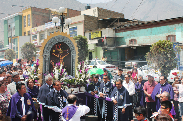 Menschen auf einer Straße mit einem Kreuz und Blumensträußen in der Mitte, fahrende Autos dahinter und Gebäude, Bäume und Straßenlaternen im Hintergrund.