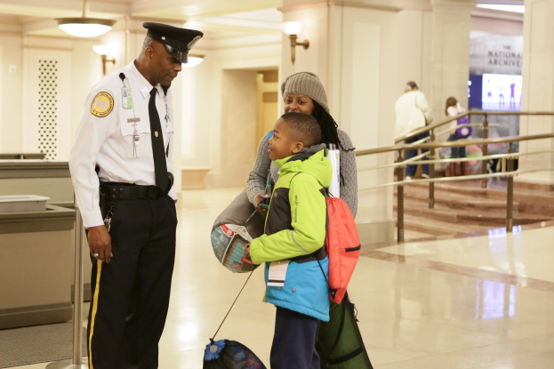 Ein Polizeibeamter in weißem Hemd, schwarzer Hose und Mütze steht neben einem jungen Jungen mit einer Tasche in einem Flughafen, mit Menschen, einem Geländer, Wandlampen und Treppen im Hintergrund.