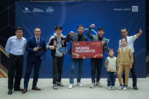 Eine Gruppe von Menschen auf einer Bühne, die eine Tafel mit dem Text "Akang Kuchaydi - Zakovat Quiz" halten, während im Hintergrund ein Banner mit Text und einem Strichcode zu sehen ist.